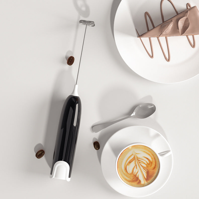 Bombom mini handheld milk frother in black next to coffee cup with latte art and spoon on white surface